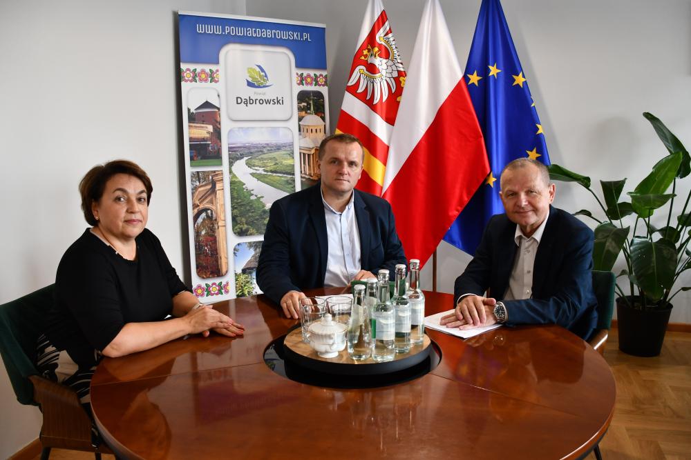Trzy osoby siedzą przy stole w biurze – kobieta i dwóch mężczyzn. Wszyscy patrzą w stronę obiektywu. . Spotkanie odbywa się w Starostwie Powiatowym w Dąbrowie Tarnowskiej.