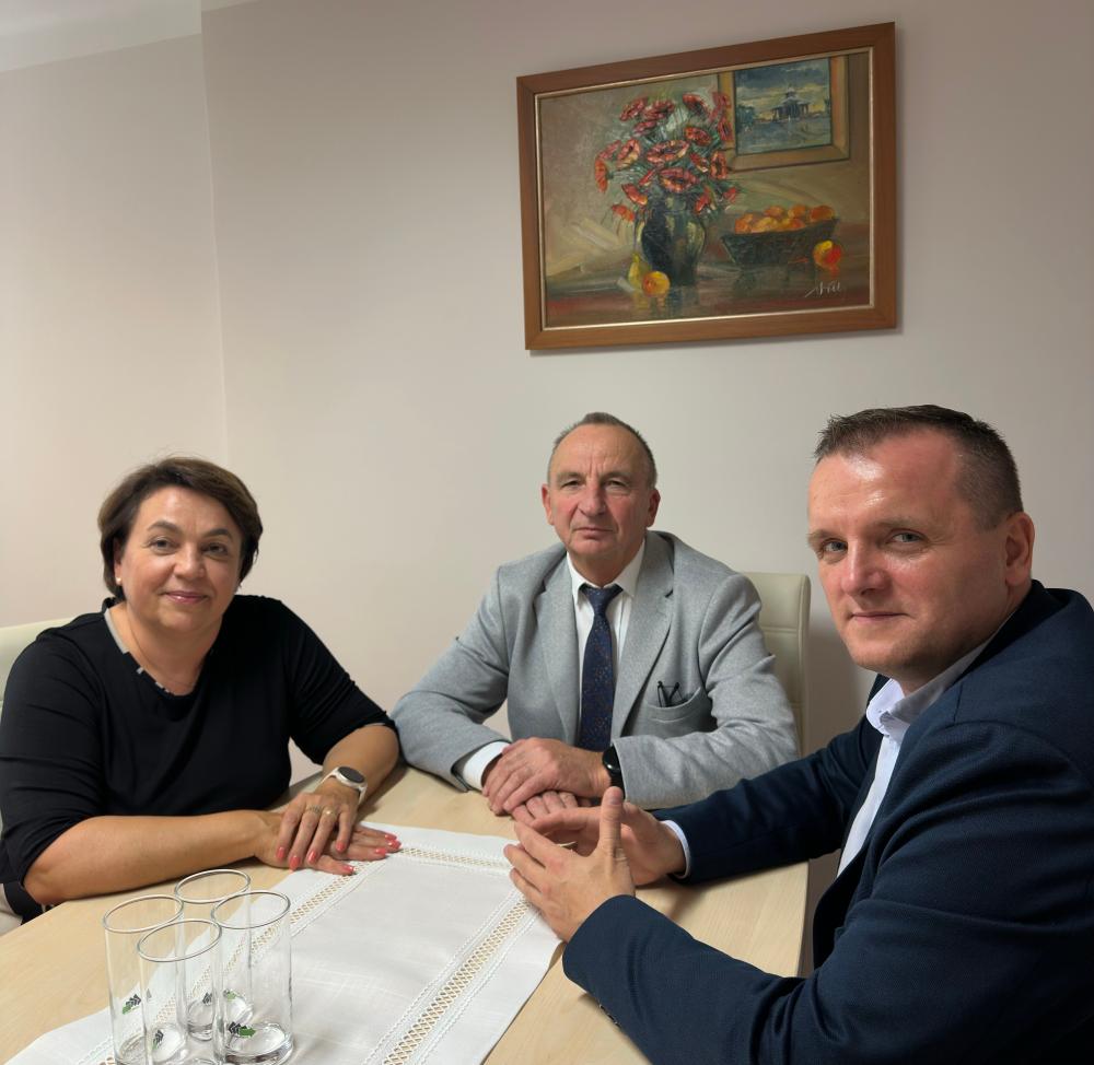 Trzy osoby siedzą przy stole w biurze – kobieta i dwóch mężczyzn. Wszyscy patrzą w stronę obiektywu. Na ścianie za nimi wisi obraz z kwiatami w wazonie. Spotkanie odbywa się w Powiatowym Urzędzie Pracy w Dąbrowie Tarnowskiej.